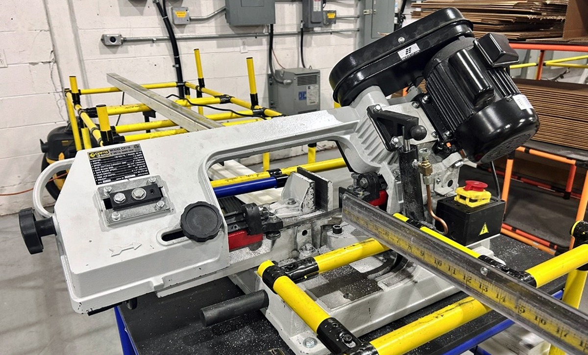 Industrial metal cutting bandsaw cutting multiple Flexpipe tubes on a yellow and blue modular workstation.