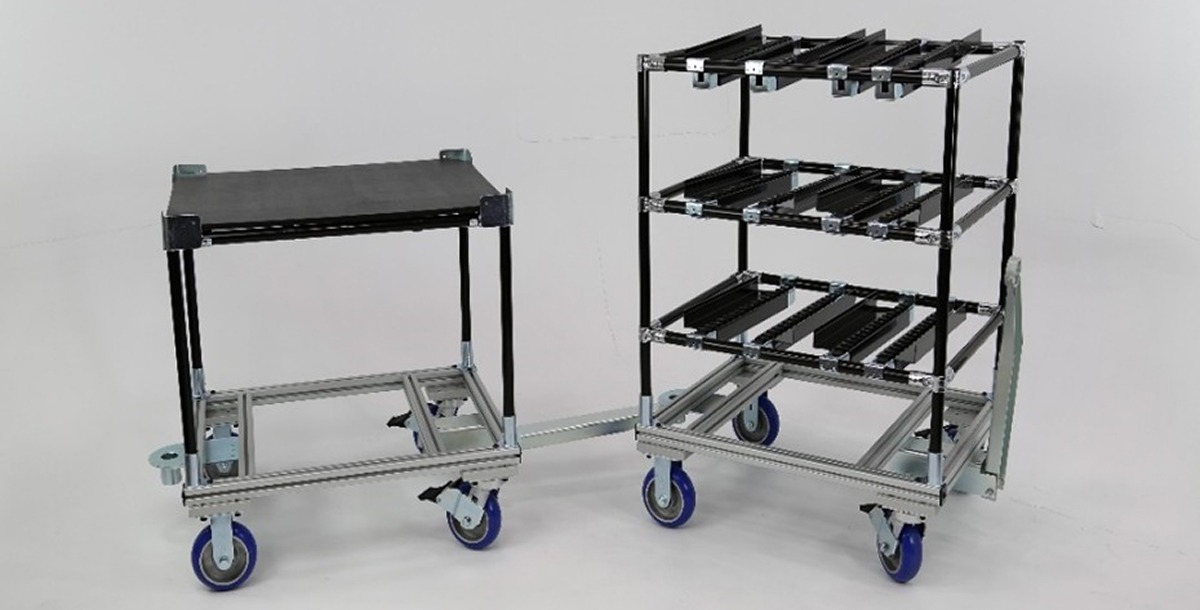 hybrid material handling setup composed of two carts linked in a mother-daughter configuration. The base structures use 80/20 aluminum extrusion, while the vertical and flow rack components are built from Flexpipe's black-coated steel tubes. One cart includes a flat working surface, and the other features three roller track flow levels for efficient kitting or parts presentation. Blue polyurethane casters and tow bar integration support tugger train logistics.