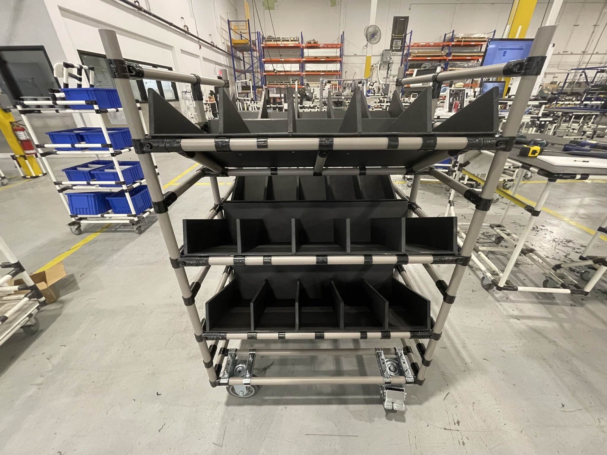 Angled industrial pipe rack with black partitioned trays, designed for part organization and kitting in a lean manufacturing or assembly line environment.