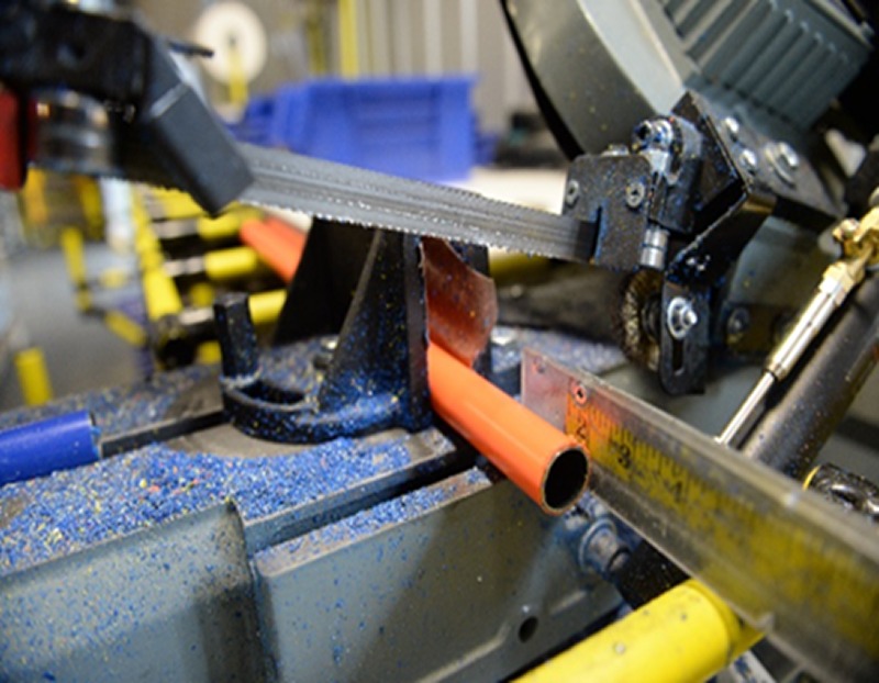 A close-up of a bandsaw cutting a coated metal tube at Flexpipe’s factory, where tubes are prepared for pre-cut services and assembly solutions. The image highlights the precision cutting process, essential for creating modular structures using Flexpipe’s tube and joint system. Metal shavings, a measuring guide, and industrial equipment emphasize the efficiency and quality control behind Flexpipe’s material handling solutions.