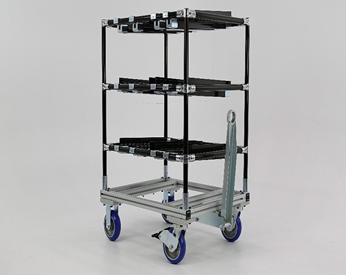 A freestanding modular tugger cart built from a combination of Flexpipe black-coated steel tubes and 80/20 aluminum extrusion. The structure includes three wide shelving levels and is mounted on heavy-duty blue casters. A metallic tow bar is installed at the front, enabling the cart to be used in a train configuration for lean material handling.