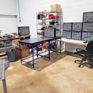Organized workspace with a blue-framed workstation, storage cabinets, and wire shelving in an electrical equipment manufacturing facility