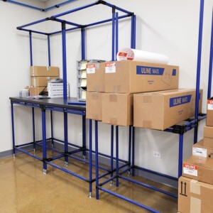 Modular blue pipe workstation with integrated storage racks and overhead shelving for electrical component handling