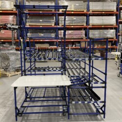 Modular blue pipe flowrack system with integrated roller tracks and a white work table in a warehouse.
